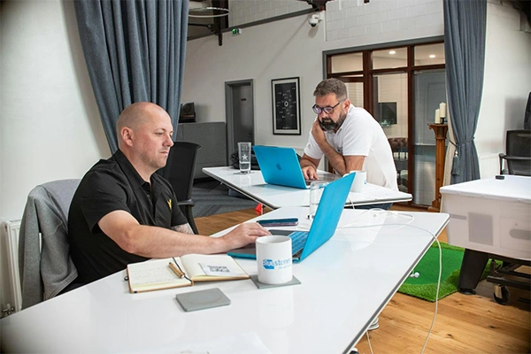 Two men working on blue laptops at white desks in a bright office